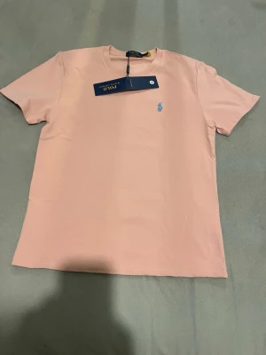 Ljusrosa t-shirt från Polo Ralph Lauren - Snygg ljusrosa t-shirt från Polo Ralph Lauren med klassisk blå logga broderad på bröstet. T-shirten har rund halsringning och korta ärmar. Tillverkad i mjuk bomull för en skön känsla och enkel stil.
