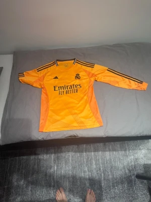 Real Madrid orange - Snygg långärmad från Real Madrid i en stark orange färg med svarta detaljer och Adidas-logga. Tröjan har klubbmärke på bröstet och 'Emirates Fly Better' tryckt framtill. Tillverkad i lätt och ventilerande polyester, perfekt för fotboll.