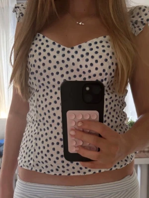 Vit prickig linnetopp med volang - Söt vit linnetopp med mörkblå prickar och volangdetaljer vid axlarna. Toppen har v-ringad halsringning och är ärmlös.