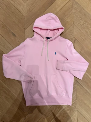Ralph Lauren hoodie - Ljusrosa hoodie med huva och dragsnören från Polo Ralph Lauren. Klassisk design med broderad blå logotyp på bröstet och stor magficka. Mjuk bomullskänsla och ribbade muddar vid ärmslut och nederkant. Perfekt för en avslappnad look.