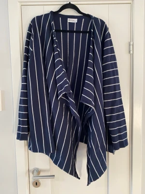 Marinblå randig kofta från Ulla Popken - Snygg marinblå kofta från Vila Joy med vita vertikala ränder. Koftan har öppen front med draperad och asymmetrisk nederkant samt långa ärmar. Tillverkad i mjukt och lätt tyg, perfekt för lager på lager och en chill look. Ny marinblå och vit randig öppen kofta strl 54/56 fr Ulla Popken  Ny, aldrig använd   Mått Över bröstet: ca 67 cm Längd från axeln och ner: ca 83 cm  70% bomull och 30% polyamid   Kommer från ett djur och rökfritt hem