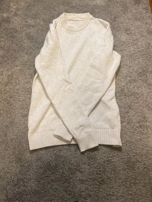 Vit stickad tröja med rund hals - En stilren vit stickad tröja med rund halsringning och ribbade muddar vid ärmslut och nederkant. Tröjan har en klassisk passform och är perfekt för lager-på-lager. Materialet känns mjukt och bekvämt mot huden.