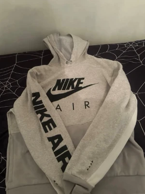 Grå Nike Air hoodie med tryck - Snygg grå Nike Air hoodie med stor svart logga på bröstet och texten 'NIKE AIR' längs ena ärmen. Hoodien har meshfoder i huvan och är tillverkad i mjukt bomullsmaterial. Perfekt för en sportig och avslappnad stil.