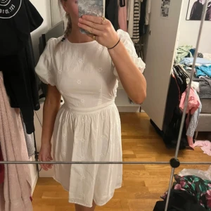 Vit kort klänning med öppen rygg - Supersöt vit kort klänning med puffärmar, broderade små blommor och öppen rygg med knyt i midjan. Perfekt för sommarens alla tillfällen och ger en romantisk vibe. Tillverkad i lätt bomullstyg som känns luftigt och bekvämt.