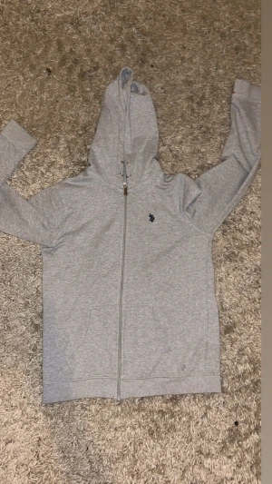 Ljusgrå hoodie från U.S. Polo Assn - Säljer en ljusgrå hoodie från U.S. Polo Assn med dragkedja framtill och två fickor. Tröjan har en broderad logga på bröstet och justerbar huva med snören. Tillverkad i mjukt bomullsmaterial som är skönt att ha på sig.