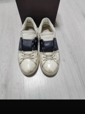 Valentino vita sneakers med blå detalj - Säljer ett par vita sneakers från Valentino med bred mörkblå läderdetalj över snörningen och nitar på sidorna. Klassisk låg modell med rund tå och platt sula. Märkeslogga på plösen. Perfekta för dig som gillar exklusiva och trendiga skor.