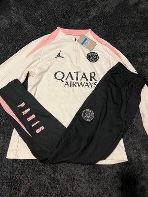 PSG tracksuit vitt och svart - Psg tracksuit med långärmad vit tröja med rosa detaljer och svart byxa. Bara att höra av er vid frågor!