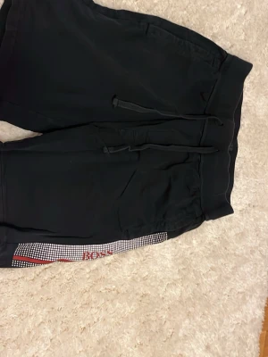 Svarta shorts från BOSS med tryck - Svarta shorts från BOSS med vit och röd grafisk detalj längs benet och logotyp. Shortsen har snörning i midjan och sidofickor. Perfekta för en avslappnad stil och tillverkade i mjukt material.