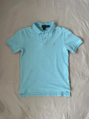 Ljusblå pikétröja från Polo Ralph Lauren - Säljer en ljusblå pikétröja från Polo Ralph Lauren med klassisk krage, knappar vid halsen och den ikoniska rosa logotypen broderad på bröstet. Tröjan har korta ärmar och är tillverkad i mjuk bomull. Perfekt för en avslappnad och stilren look.