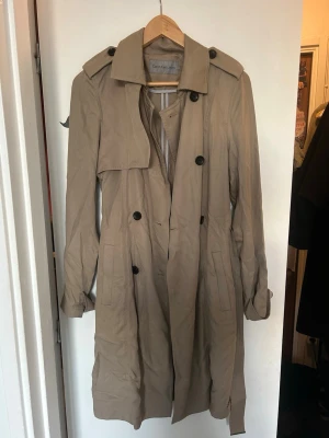 Beige trenchcoat från Calvin Klein - Stilren beige trenchcoat från Calvin Klein. Jackan har långa ärmar, fickor framtill och detaljer som knappar vid ärmslut. Perfekt för dig som vill ha en tidlös och trendig look.