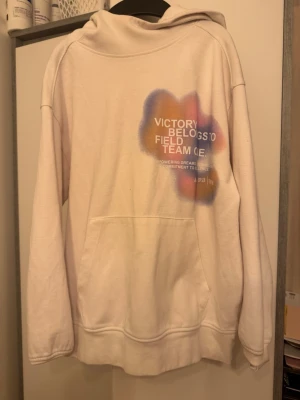 Vit hoodie med färgstänk och tryck - Säljer en vit hoodie från Adidas med färgstänk i blått, orange och lila samt tryck på både fram- och baksida. Texten 'Victory belongs to field team one' syns tydligt. Hoodien har stor ficka framtill och klassisk huva.