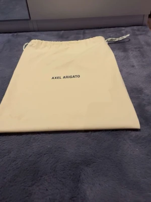 Beige dustbag från Axel Arigato - Säljer en beige dustbag från Axel Arigato med dragsko upptill. Perfekt för att förvara skor eller accessoarer. Dustbagen är i ett mjukt tygmaterial och har enkel, stilren design med logga på framsidan.