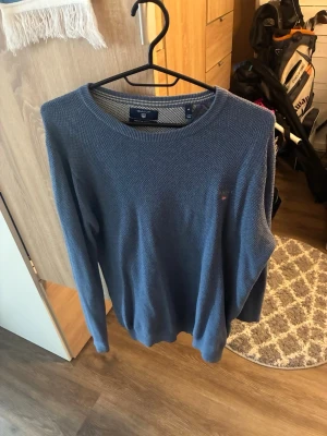 Blå stickad tröja från GANT - Snygg blå stickad tröja från GANT i premium bomull. Tröjan har rund halsringning, diskret GANT-logo broderad på bröstet och ribbade muddar vid ärmslut och nederkant. Perfekt för en stilren och avslappnad look.