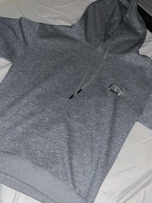 Grå hoodie  - Snygg grå hoodie från med korta ärmar och broderad logga på bröstet. Hoodien har en klassisk huva med dragsko och en stor magficka framtill. Tillverkad i mjukt bomullsmaterial som känns skönt mot huden. Liten i storlek!!