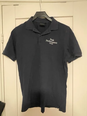 Marinblå Peak Performance piké - Marinblå pikétröja från Peak Performance med klassisk krage och korta ärmar. Diskret broderad logga i vitt på bröstet och knappar vid halsen. Tillverkad i mjuk bomull för en clean och sportig look.