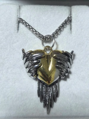 Halsband med hjärta och vingar - Unikt halsband med ett guldigt hjärta omslutet av detaljerade silverfärgade vingar. Hänget har även fjäderliknande detaljer som hänger ner, vilket ger en cool och edgy look, 4,3 x 3 cm. Kedjan är i silverfärg och har en klassisk länkdesign, 60 cm. Perfekt för dig som gillar statement-smycken.