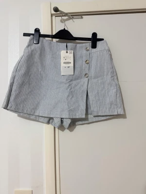 Randig skort från Zara  - Snygg skort från Zara i vitt och ljusblått med glitter och tunna vertikala ränder. Modellen har hög midja, dekorativa knappar framtill och en omlott-look. perfekt för sommaren. Klassisk och trendig design.