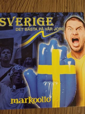 CD Markoolio - Sverige Det Bästa På Vår Jord - CD-skiva med Markoolios låt 'Sverige Det Bästa På Vår Jord'. Omslaget har blå och gula färger med en stor uppblåsbar hand i svensk flaggdesign. Perfekt för dig som gillar svensk musik och vill ha en samlarpryl med landslagstema.