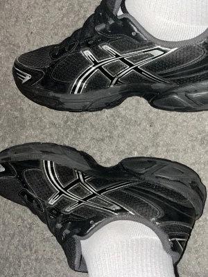 Svarta Asics sneakers med silverdetaljer - Säljer ett par svarta Asics sneakers med snygga silverdetaljer och meshpaneler för bra ventilation. Skorna har en sportig look med låg profil och robust sula som passar perfekt till streetwear-stilen. Klassisk Asics-logga på sidan och snörning framtill. Får även med ordinarie box. 
