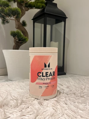 MyProtein Clear Whey Protein Peach Tea - Mycket mindre än halva kvar ( liten portion ) Säljer en burk MyProtein Clear Whey Protein i smaken Peach Tea. Förpackningen är vit med rosa och persikofärgade detaljer och tydlig text på framsidan. Perfekt för dig som vill ha ett fräscht proteintillskott.