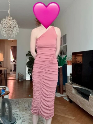 Rosa oneshoulder klänning med scrunch - Säljer en rosa oneshoulder klänning med rynkade detaljer längs hela framsidan och baksidan. Klänningen är figurnära och har en längre, smal siluett som slutar vid vaden. Perfekt för bröllop, fest eller nu till våren och sommaren. Väldigt bekväm och känns lyxig och exklusiv. Jag är 170 cm för referens. Passar en S eller XS. Oanvänd. Har stretch och är väldigt bekväm.
