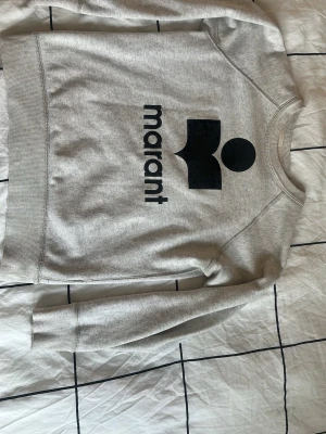 Grå sweatshirt från Isabel Marant - Snygg grå sweatshirt från Isabel Marant med stort svart logotryck på bröstet. Som också är ganska utsuddat. Tröjan har rund hals, långa ärmar och ribbade muddar vid ärmslut och nederkant. Tillverkad i mjukt bomullsmaterial som är skönt mot huden. Den är i storlek 36 som jag skulle säga motsvarar storlek XS eller S. Den har också en liten permanent fläck på ryggen som inte syns. Det är typ en liten prick. 