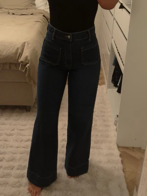 Mörkblå utsvängda jeans med hög midja - Säljer ett par mörkblå jeans med hög midja och vida ben. Jeansen har stora framfickor och markerade sömmar för en cool retrostil. Tillverkade i kraftigt denimtyg som ger en snygg siluett. Perfekta för dig som gillar trendiga och bekväma jeans.