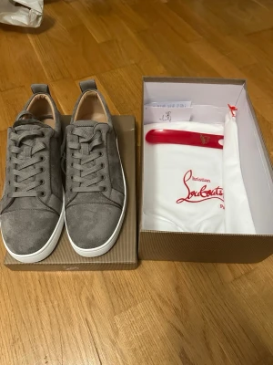 Gråa sneakers från Christian Louboutin - Snygga gråa sneakers från Christian Louboutin i mocka med vita sulor och matchande grå skosnören. Klassisk låg modell med diskret logga på sidan och stilren design. Perfekta för dig som vill ha något exklusivt och trendigt.