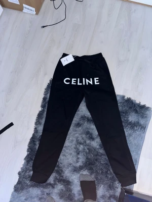 Svarta mjukisbyxor från Celine - Snygga svarta mjukisbyxor från Celine med stort vitt logotryck framtill. Byxorna har resår i midjan med dragsko och muddar vid bensluten. Perfekta för en chill och trendig look. Tillverkade i mjukt material för maximal komfort.