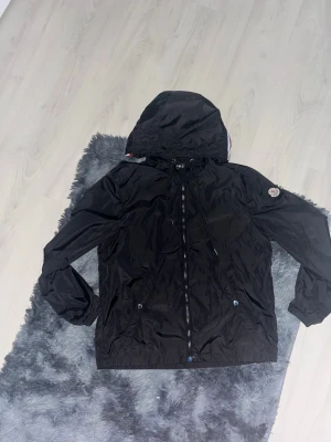 Svart vindjacka från Moncler - Snygg svart vindjacka från Moncler med huva och dragkedja framtill. Jackan har Moncler-logga på ärmen, två fickor med tryckknappar och en vit-röd-blå rand på huvan. Tillverkad i lätt, glansigt material som står emot vind.