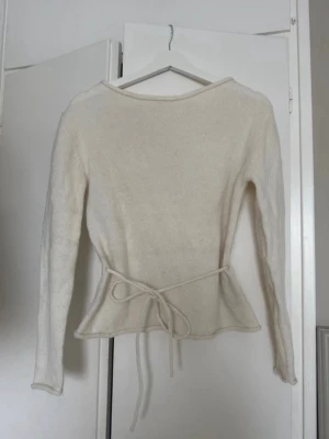 Vit stickad tröja med knytband - Säljer en krämvit stickad tröja med långa ärmar och ett snyggt knytband i midjan som ger en feminin siluett. Tröjan har rund halsringning och lätt utsvängd nederkant. Perfekt för lager-på-lager och enkel att styla till olika outfits. Materialet är från alpacka