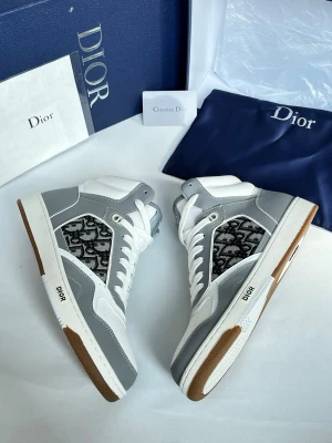 Dior gråa och vita high-top sneakers - Exklusiva Dior high-top sneakers i vitt och grått läder med ikoniskt Dior-monogram i textil på sidan. Skorna har vita snören, gummisula med brun detalj och tydlig Dior-logga på sulan. Perfekt för dig som vill sticka ut med lyxig streetstyle.
