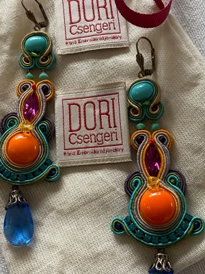 Färgglada örhängen från Dori Csengeri från Svensk Tenn. Hand made. - Unika statementörhängen från Dori Csengeri med broderade detaljer i turkos, orange, lila och gult. Stora orange pärlor, lila stenar och blå droppformade hängen. Handgjorda och riktigt iögonfallande accessoarer för dig som gillar färg och kreativ design.