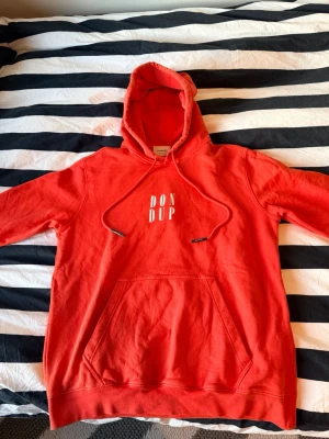 Röd hoodie från Dondup med tryck - Snygg röd hoodie från Dondup med vit logotyp på bröstet som ger en tydlig streetwear-look. Klassisk känguruficka framtill, justerbar dragsko i huvan och mjukt bomullsmaterial för hög komfort. Perfekt för dig som vill sticka ut med en färgstark och stilren outfit.  ✔ 100% äkta ✔ Mjuk och bekväm ✔ Klassisk streetwear-design ✔ Färgstark och trendig look