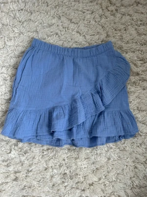 Blå volangkjol  - Söt blå kortkjol med volangdetaljer och omlottkänsla. Kjolen har resår i midjan och är gjord i ett mjukt, luftigt bomullstyg. Passar mog som vanligtvis har XS/S💕