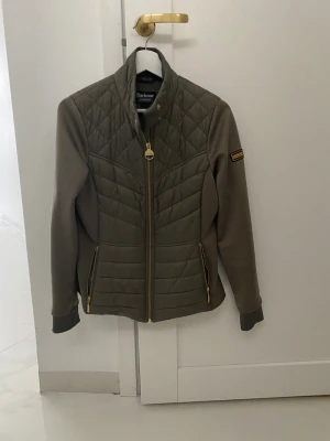 Olivgrön quiltad jacka från Barbour - Snygg olivgrön jacka från Barbour med quiltade partier och guldfärgade dragkedjor. Jackan har hög krage, lång ärm och två fickor framtill. Perfekt för dig som gillar klassisk men modern stil.
