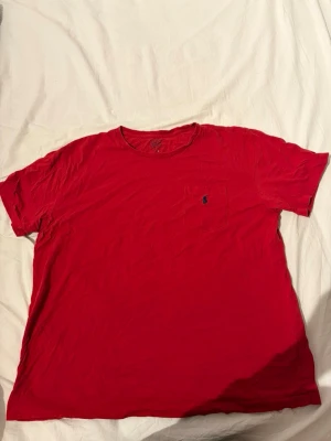 Röd t-shirt från Polo Ralph Lauren - Klassisk röd t-shirt från Polo Ralph Lauren med rund halsringning och korta ärmar. Bröstficka med den ikoniska blå broderade loggan. Tillverkad i mjuk bomull för en skön känsla. Perfekt till jeans eller shorts för en enkel och stilren look.
