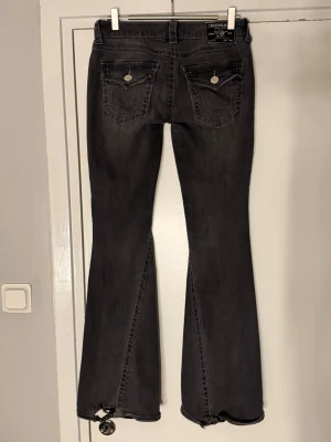 Svarta joey low rise flare jeans - True Religion - Säljer ett par svarta flare jeans från True Religion med klassiska kontrastsömmar och snygga fickdetaljer bak. Jeansen har låg midja. Materialet är denim med stretch. Lite hål längst ner bak på jeansen som syns på bild!
