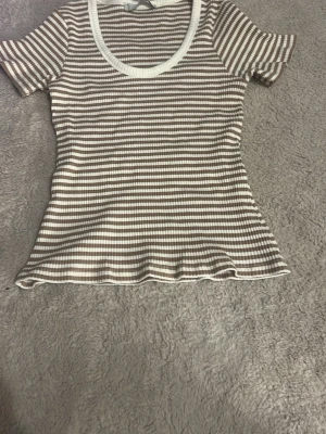 Randig ribbad t-shirt med rund hals - Snygg ribbad t-shirt med beige och vita horisontella ränder och rund, vit kantad halsringning. T-shirten har korta ärmar och en figurnära passform som ger en fräsch och trendig look. Perfekt att matcha med jeans eller kjol.