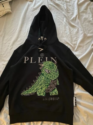 Svart hoodie Plein med Godzilla-motiv - Svart hoodie från Philipp Plein med stor huva och snörning. Framsidan pryds av ett färgglatt Godzilla-motiv i grönt och lila, dekorerat med glittrande stenar och texten 'PLEIN' samt 'LIL GOD$ILLA'. Perfekt för dig som gillar statement-plagg och streetwear.