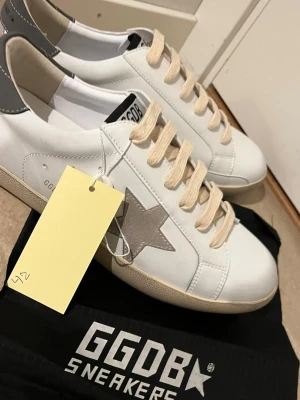 Golden Goose vita sneakers stjärna - Snygga vita sneakers från Golden Goose med grå mockadetaljer, beige skosnören och den ikoniska stjärnan på sidan. Skorna har en låg silhuett och en vintage-inspirerad sula. Perfekta för dig som gillar streetstyle och exklusiva detaljer.