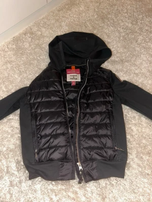 Svart pufferjacka från Parajumpers - Snygg svart pufferjacka från Parajumpers med huva och dragkedja framtill. Jackan har quiltad design på framsidan och mjuka ärmar i fleecematerial. Märkespatch på ärmen och två fickor med dragkedja. Perfekt för våren. Använd max 5 ggr.