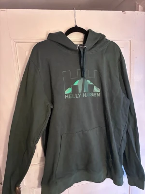 Mörkgrön Helly Hansen hoodie - Mörkgrön hoodie från Helly Hansen med stor logga och bergmotiv på bröstet. Klassisk känguruficka framtill och justerbar huva med dragsko. Tillverkad i mjukt bomullsmaterial, perfekt för chill dagar. Regular fit. Passar snarare S/M