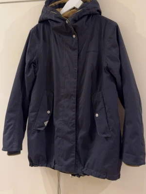 Mörkblå parkas från Didriksons - Mörkblå parkasjacka från Didriksons med huva och två stora fickor framtill med tryckknappar. Jackan har dragsko i midjan och nederkant för justerbar passform samt diskret logga på bröstet. Perfekt för kyliga dagar och enkel att matcha.