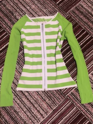 Grön och vit randig ziptröja - Snygg långärmad ziptröja med gröna och vita horisontella ränder på kroppen och helgröna ärmar. Dragkedja framtill och figurnära passform. 