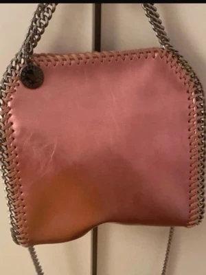 Rosa Stella McCartney handväska - Säljer en snygg rosa handväska från Stella McCartney med kedjehandtag och dekorativa sömmar längs kanterna. Väskan har en mjuk, fyrkantig form och är gjord i veganskt läder. Metallbricka med logga framtill. Perfekt för dig som vill ha en trendig accessoar.