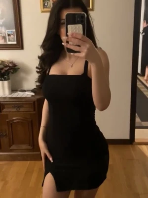 Svart kort klänning med slits - Säljer en svart, glittrig syns ej på bilden, tight kort klänning med smala axelband och en liten slits framtill. Klänningen har en enkel och stilren design som passar perfekt till olika tillfällen. Materialet känns mjukt och följsamt mot kroppen.