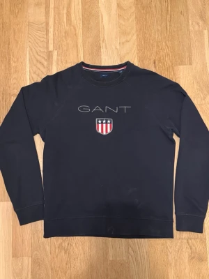 Svart sweatshirt från GANT - Klassisk svart sweatshirt från GANT med rund halsringning och broderad logga samt sköld med stjärnor och ränder på bröstet. Tröjan har långa ärmar och en relaxed passform. Perfekt för en stilren och avslappnad look.