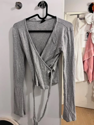 Grå omlottopp med knytband - Snygg grå omlottopp med långa ärmar och knytband i midjan. Toppen har en djup v-ringning och är tillverkad i mjuk bomullsmix som känns skön mot huden. Perfekt att styla med jeans eller kjol för en trendig look.