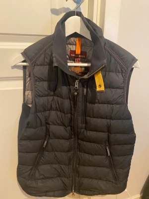 Svart dunväst från Parajumpers - Snygg svart dunväst från Parajumpers med hög krage, gul detalj vid dragkedjan och två framfickor med dragkedja. Västen har quiltad design och klassisk logga på nacken. Perfekt för lager-på-lager och håller dig varm under kyliga dagar